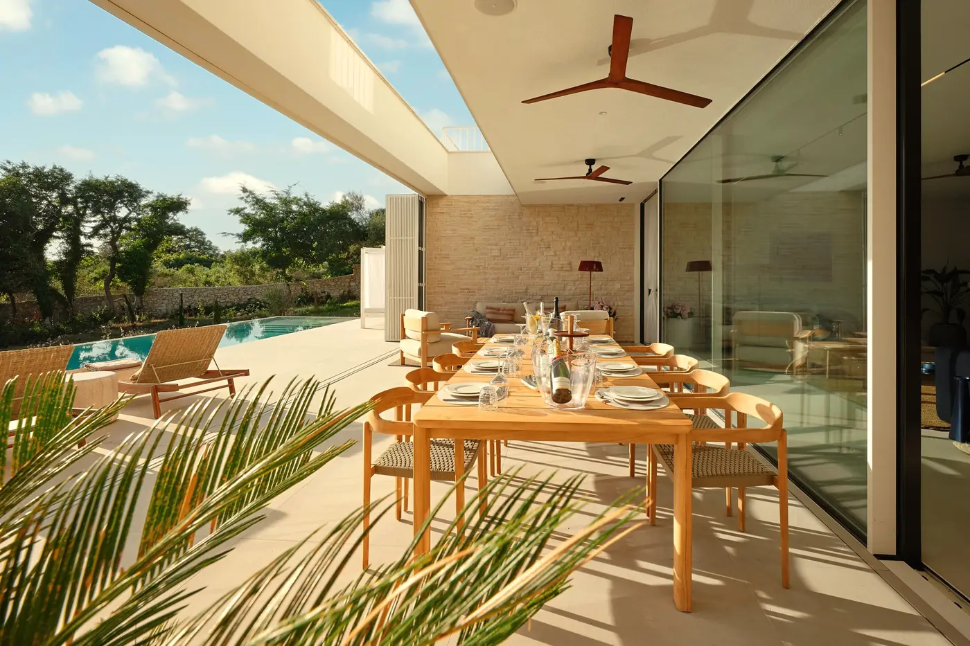 The covered terrace in daylight: a long teak table, palm shade, the pool beyond